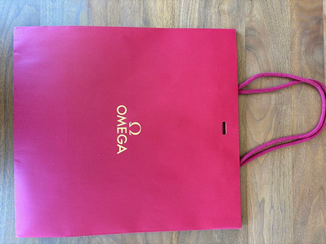 Red OMEGA paper shopping bag with gold logo and rope handles on a wooden surface.