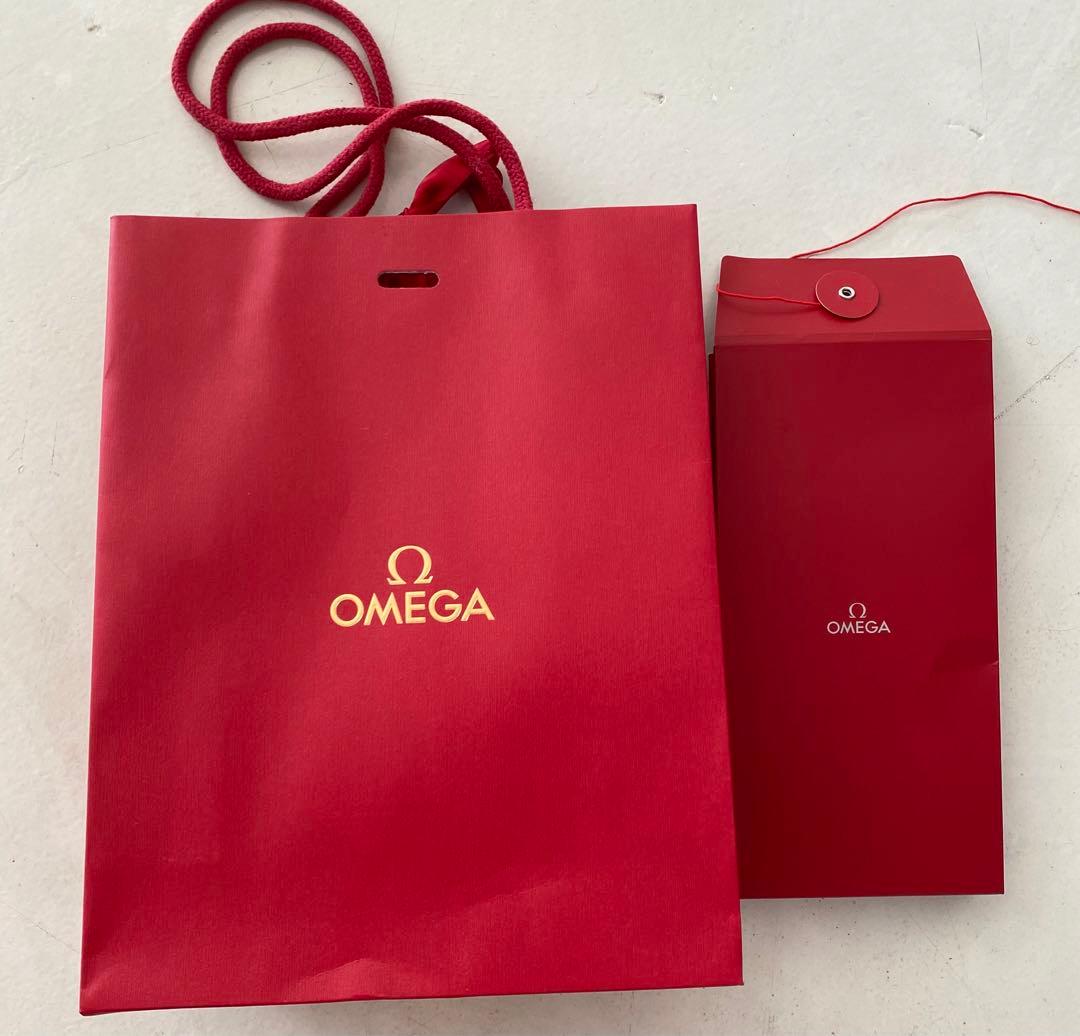 Red Omega paper shopping bag and a matching red gift envelope, both showing the Omega logo, on a white surface.