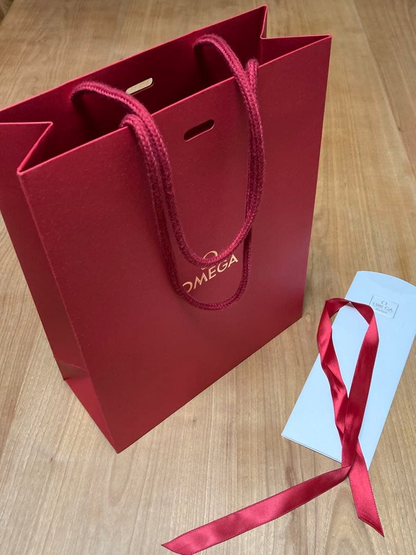 Red Omega paper shopping bag standing upright on a wooden surface with a detached red ribbon and a white tag beside it.