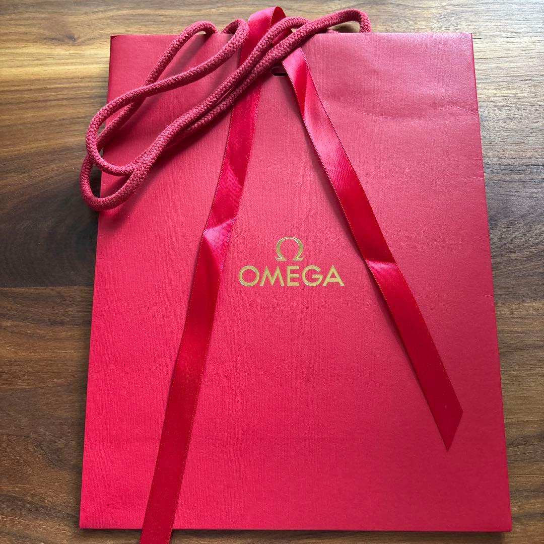 A red OMEGA paper shopping bag with a gold OMEGA logo, red ribbon, and red rope handles on a wooden surface.
