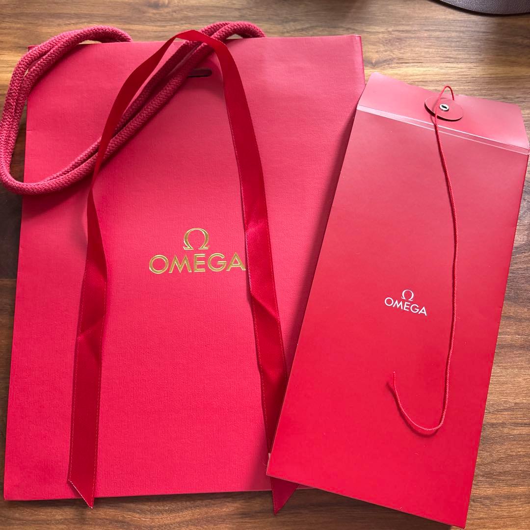 Red OMEGA paper shopping bag and a red envelope-style bag laid on a wooden surface.