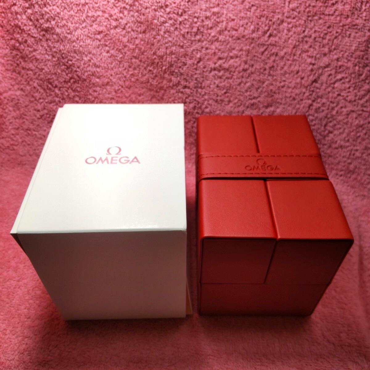 A white OMEGA outer box beside a red OMEGA watch case placed on a pink towel.