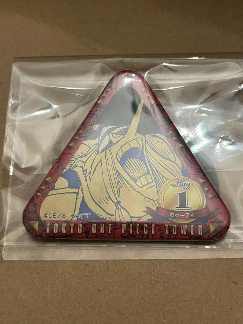 Front of a triangular One Piece Tokyo Tower limited can badge showing a character illustration and 'TOKYO ONE PIECE TOWER' text in plastic packaging.
