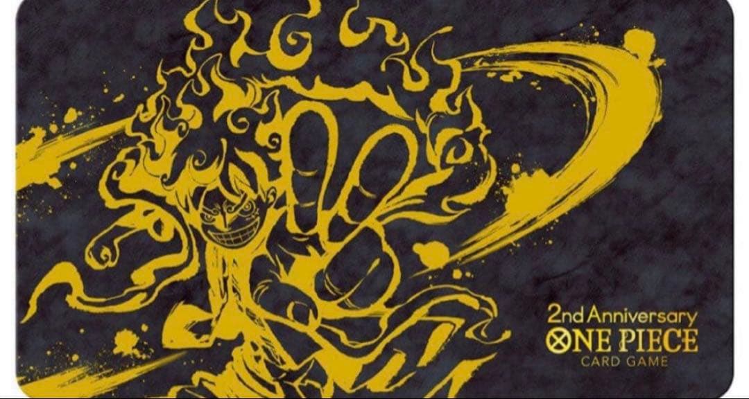 Flat polyester playmat for the One Piece Card Game with a gold flame-style illustration and "2nd Anniversary ONE PIECE CARD GAME" text on a dark background.