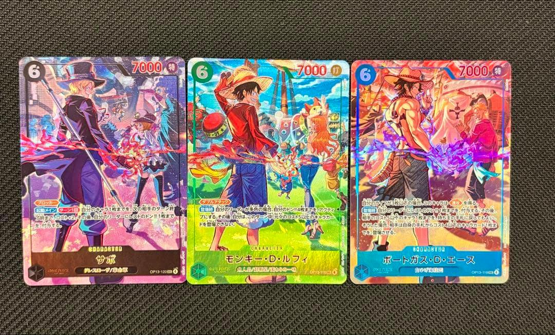 Three One Piece trading cards (ルフィ, エース, サボ) showing their front artwork arranged side-by-side on a textured dark surface.