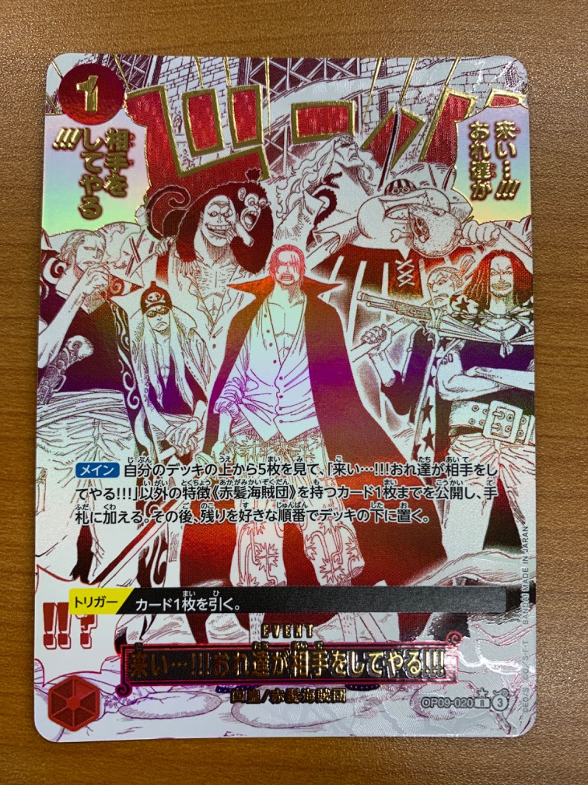 Photograph of the ONE PIECE trading card 状態A 来い…!!!おれ達が相手をしてやる!!! showing holographic foil, character artwork, and Japanese game text on a wooden surface.