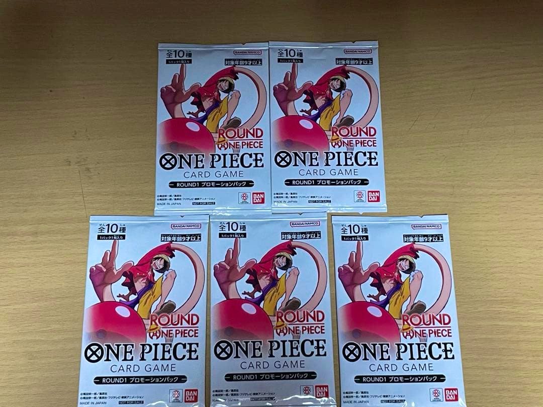 Five sealed ワンピースカードゲーム ROUND1 プロモーションパック booster packs arranged on a wooden surface.