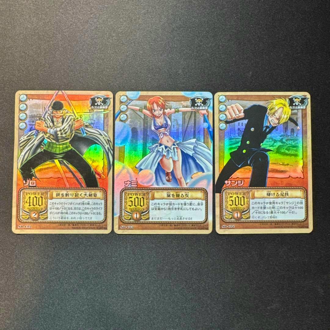 Three holographic One Piece trading cards showing Zoro, Nami, and Sanji placed side by side on a dark background.