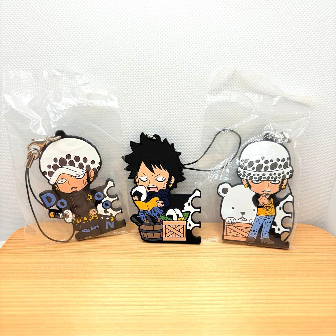 Three Trafalgar Law rubber strap keychains are displayed side-by-side, two in plastic sleeves and one unwrapped, on a wooden surface.
