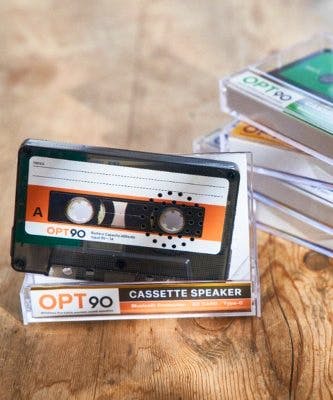 Cassette-shaped Bluetooth speaker labeled OPT90 shown upright on its clear plastic case with several cassette-style cases stacked behind on a wooden surface.