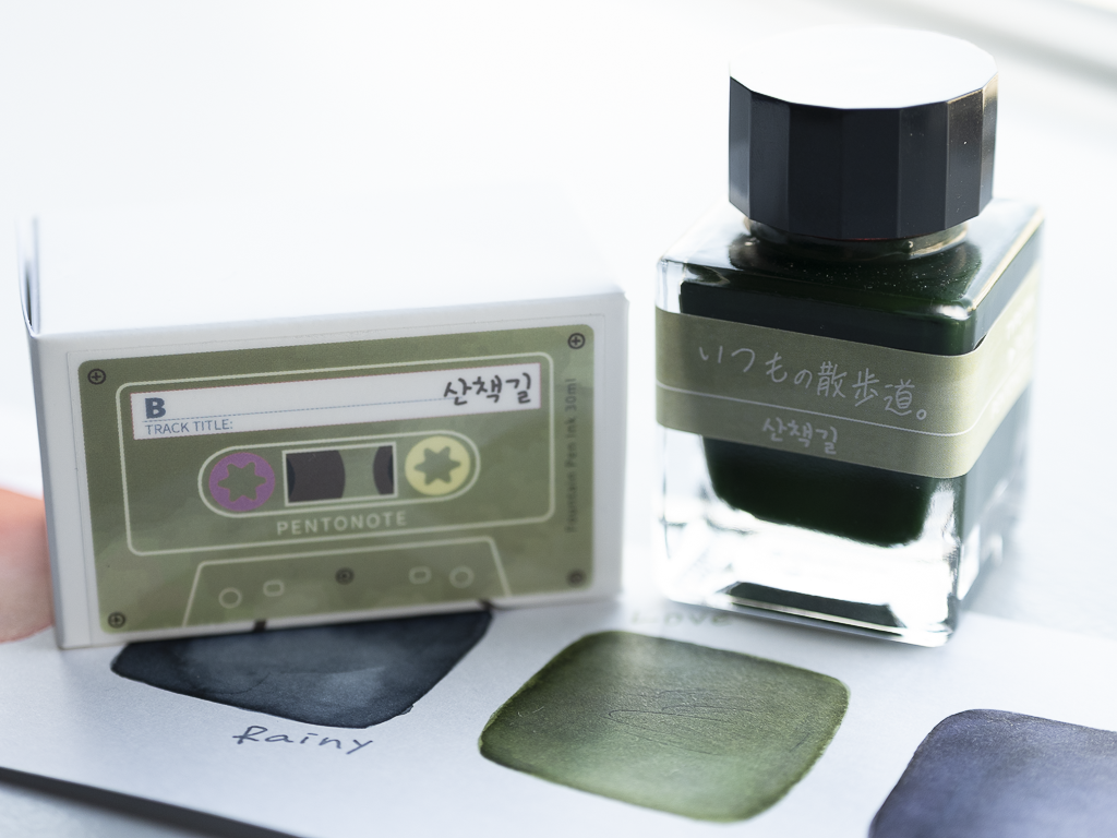 A square glass bottle of fountain pen ink next to its cassette-themed box with three ink swatches on paper.