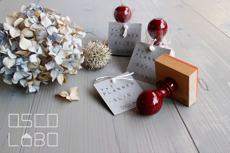 Three wooden planner stamps with red handles and paper tags arranged on a gray wooden surface with dried flowers nearby.