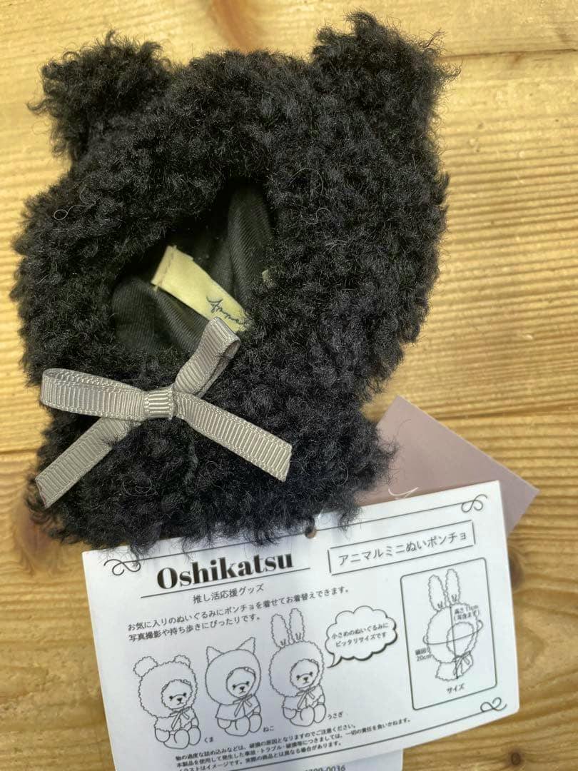 Black fuzzy bear-eared doll poncho with a gray ribbon bow and attached product tag showing illustrations.