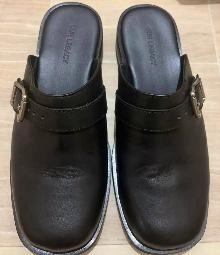 Top-down photo of a pair of OURLEGACY Camion black mule shoes shown side-by-side on a tiled floor.