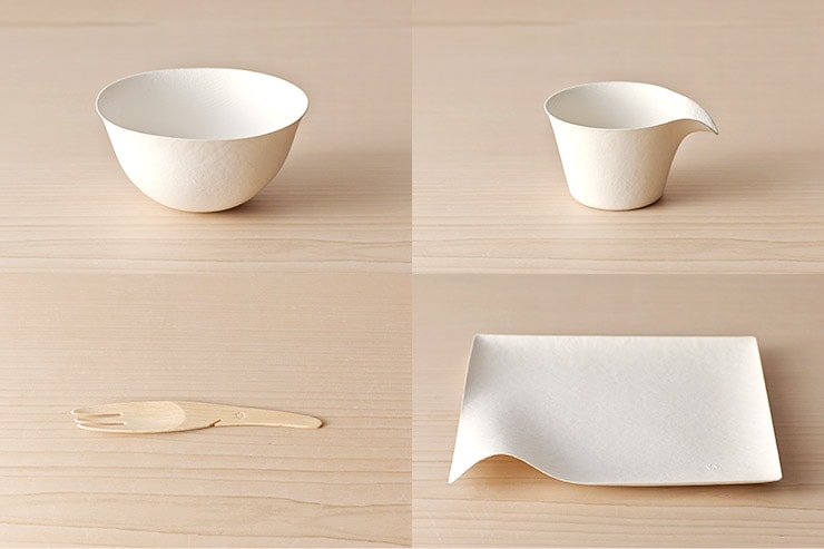 A paper outdoor tableware set on a wooden surface: a paper bowl, a paper cup, a small paper plate, and a wooden fork.