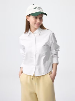 Model wearing a white Oxford short women's shirt, shown front view with hands in pockets.