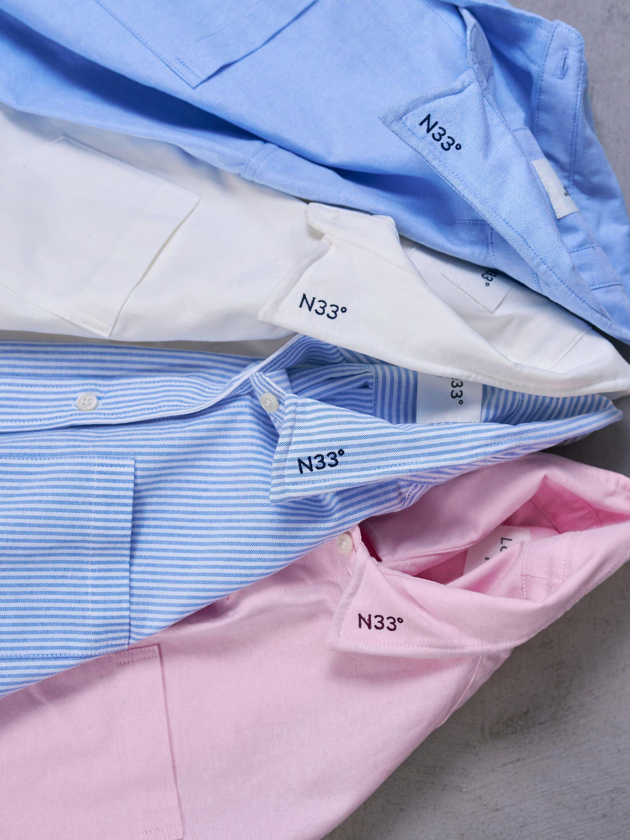 Folded men's oxford cotton shirts in blue, white, striped and pink showing collars with 'N33°' embroidery.