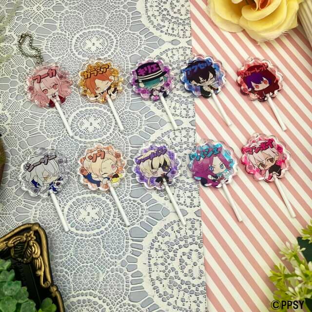 Photograph of multiple OZMAFIA!! acrylic candy-shaped keychain charms with chibi character artwork laid out on lace and striped backgrounds.