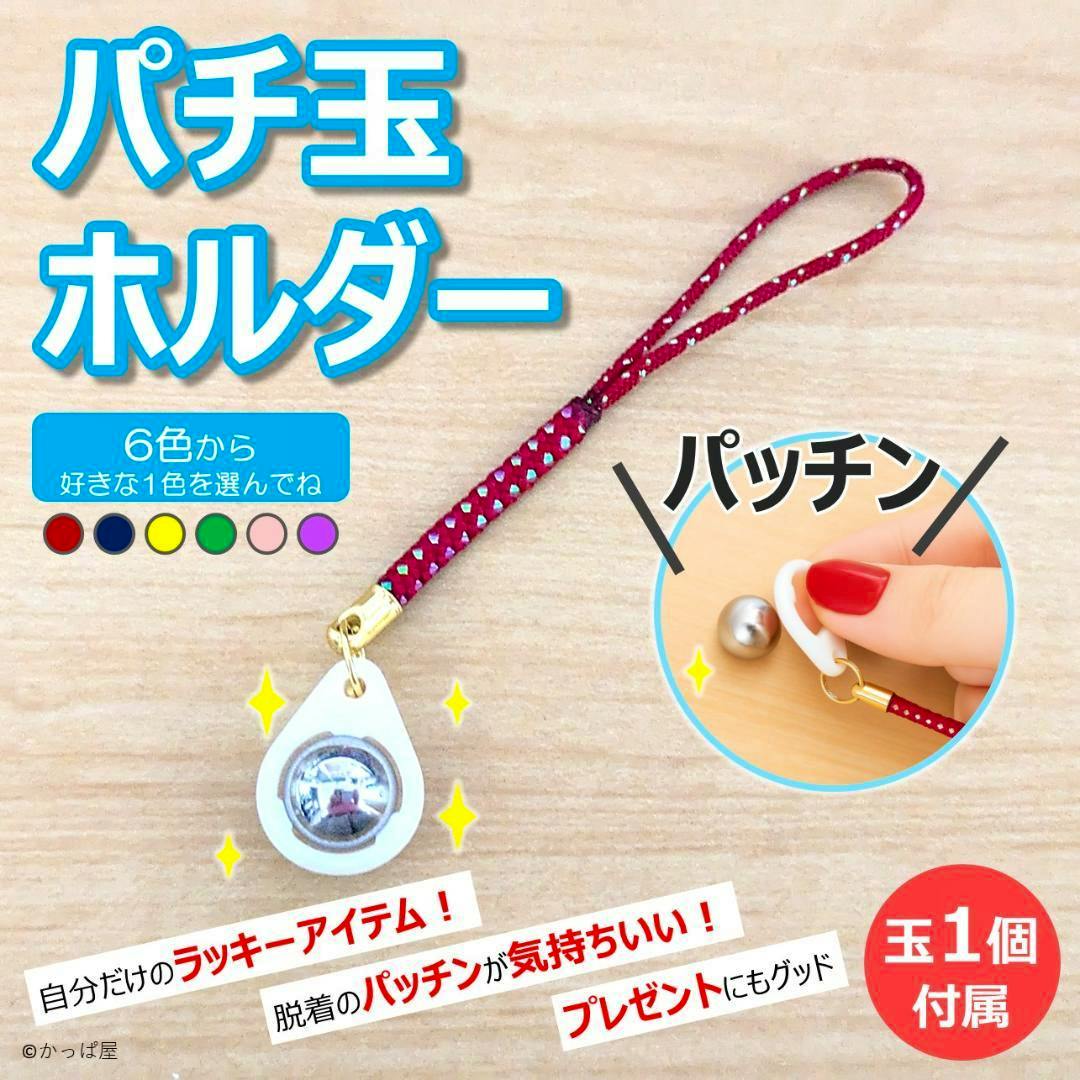 A red strap keychain with a white teardrop plastic holder containing one metal pachinko ball on a wooden surface, with an inset showing a hand removing the ball.