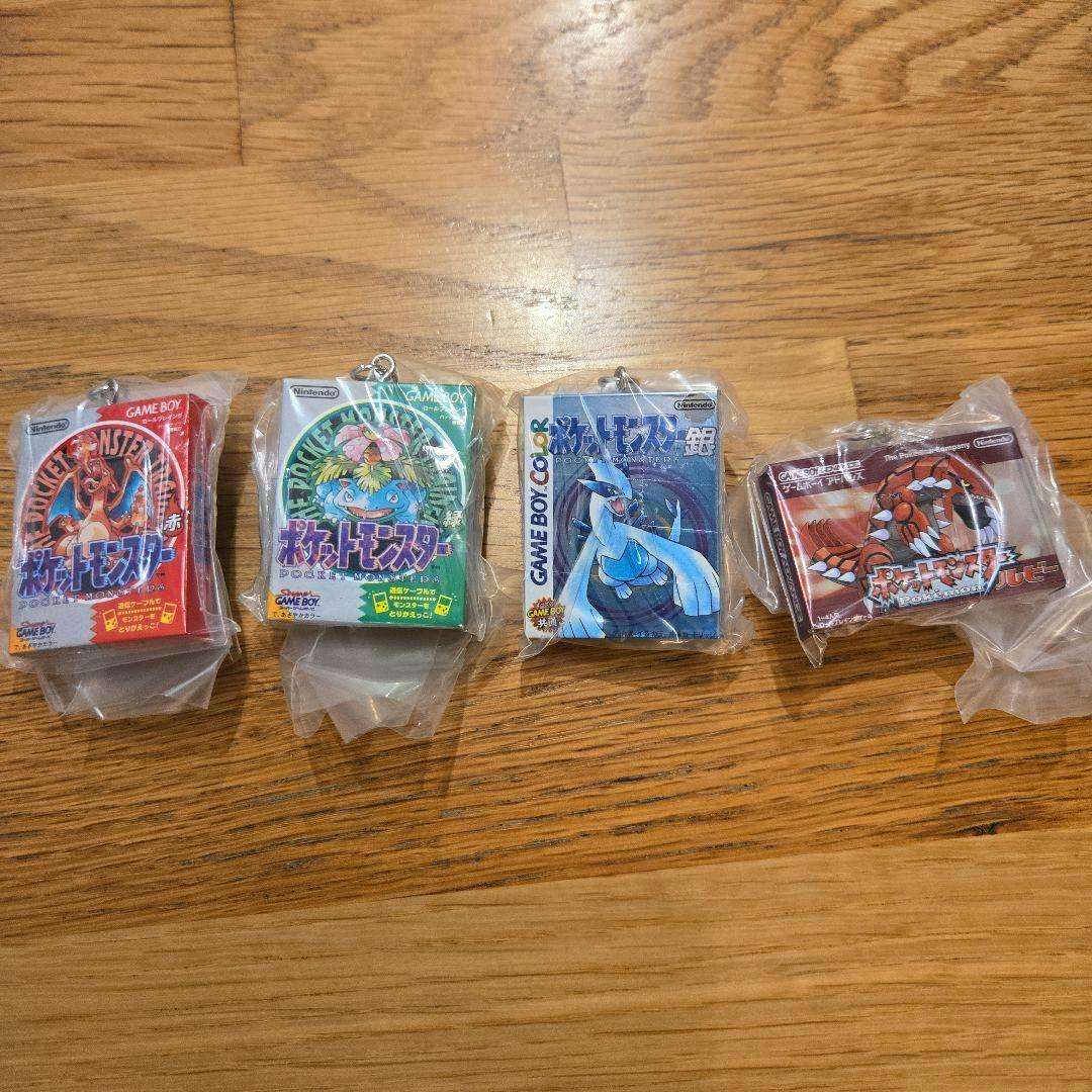Four packaged acrylic keychain charms resembling Game Boy cartridge packages laid in a row on a wooden surface.