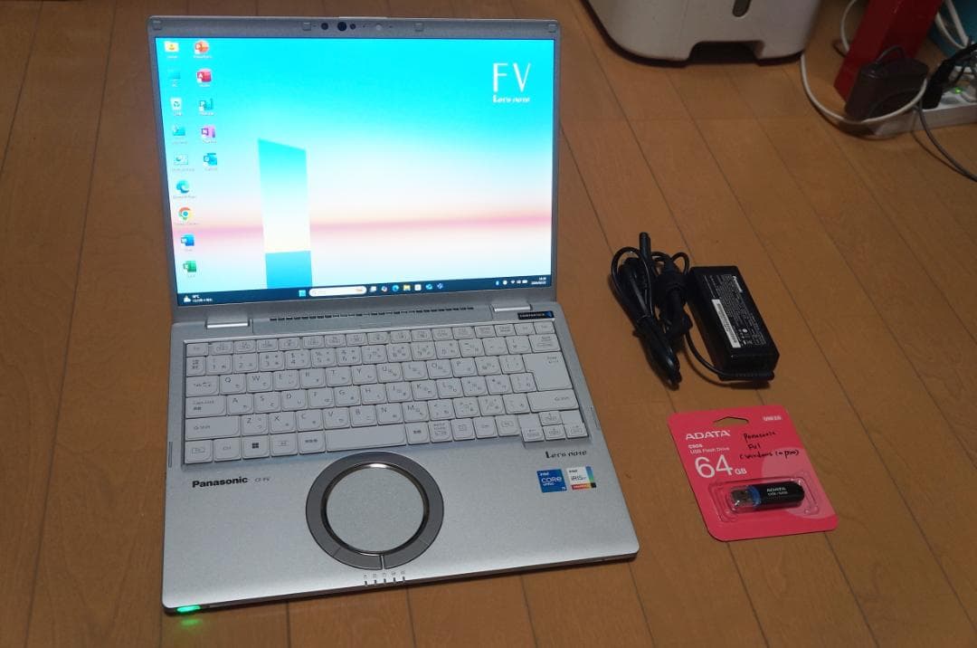 Open Let's note FV1 laptop with screen on, keyboard, power adapter, and a packaged 64GB USB drive on a wooden floor.