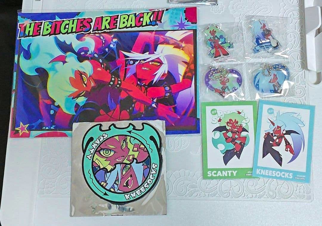 Photo showing acrylic keychain charms, stickers, and printed cards laid out on a surface with small packaged keychains visible at top right.