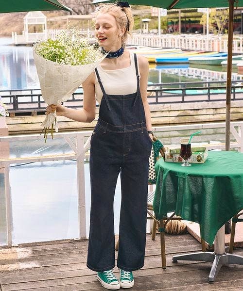 Model wearing a black denim salopette (all-in-one) with a white tank top and green sneakers, standing on a dock while holding a bouquet of flowers.