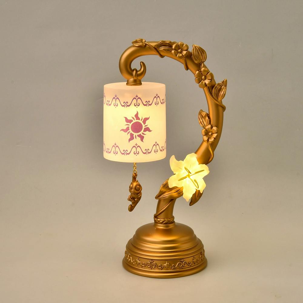 Pascal LED light 'FEEL LIKE RAPUNZEL' decorative gold table lamp with a patterned cylindrical shade, floral stem, and illuminated flower accent.