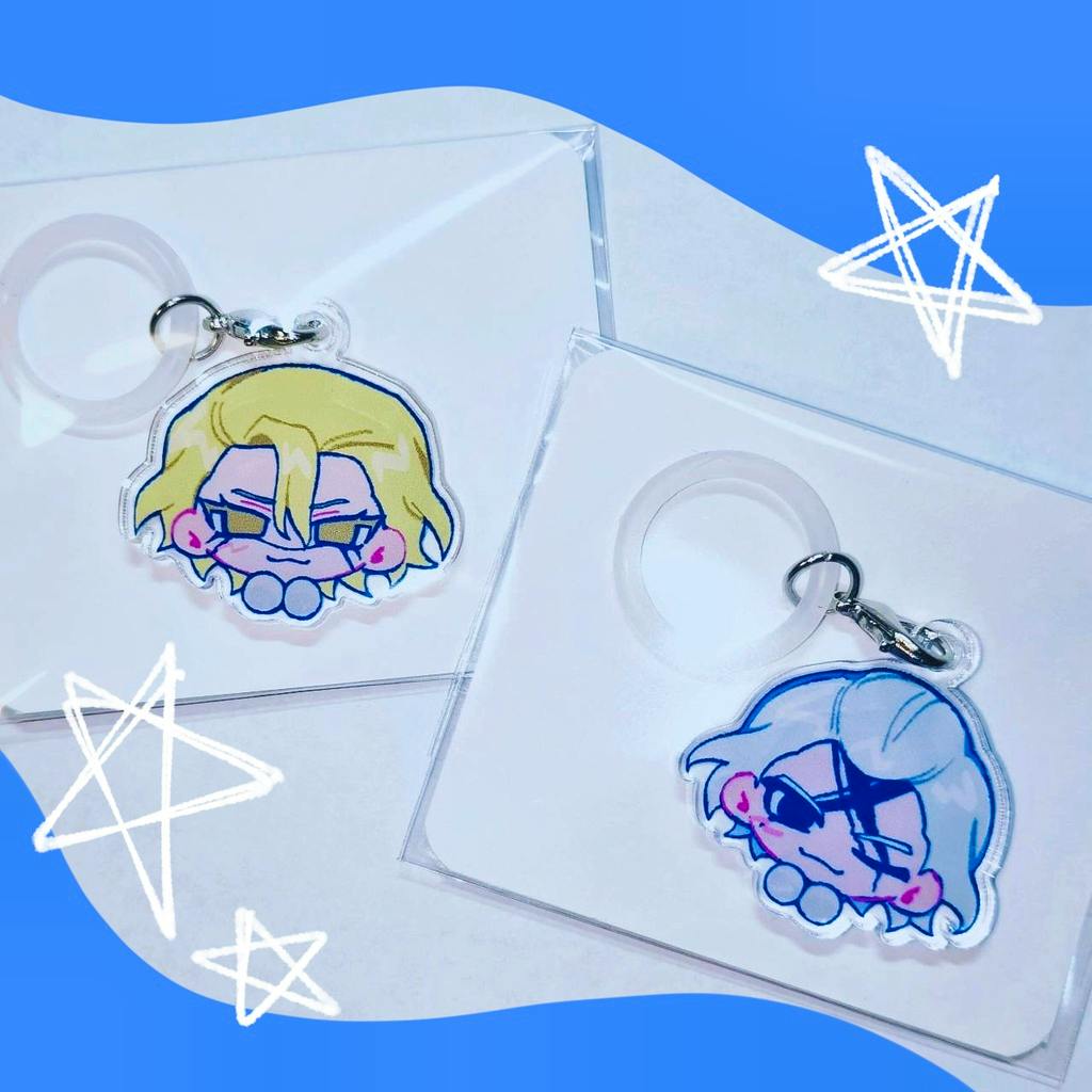 Two pastel acrylic charms with stylized face designs attached to key rings, shown on clear backing cards against a blue background.