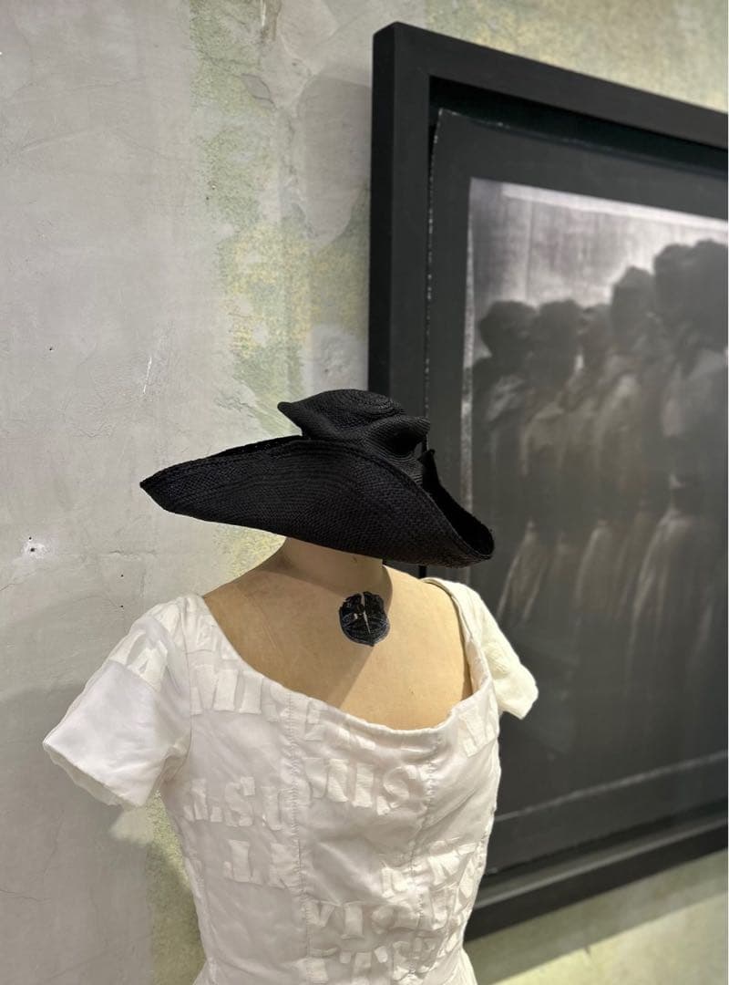 Black straw hat displayed on a dress mannequin wearing a white dress, with a framed photograph on the wall behind.