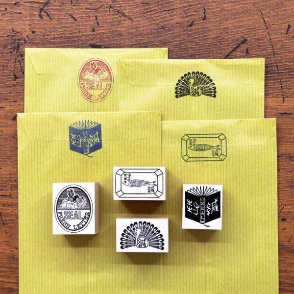 Four wooden stamps showing black printed designs, including a peacock, arranged on yellow envelopes with their stamped impressions visible.