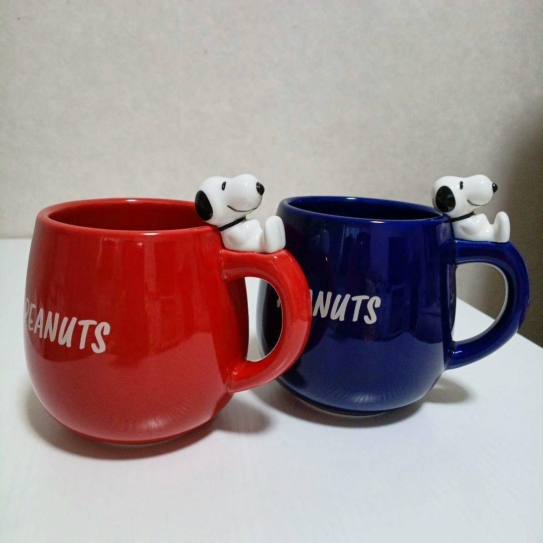 Two ceramic PEANUTS Snoopy mugs, one red and one blue, each with a small Snoopy figure attached to the handle, shown side by side.