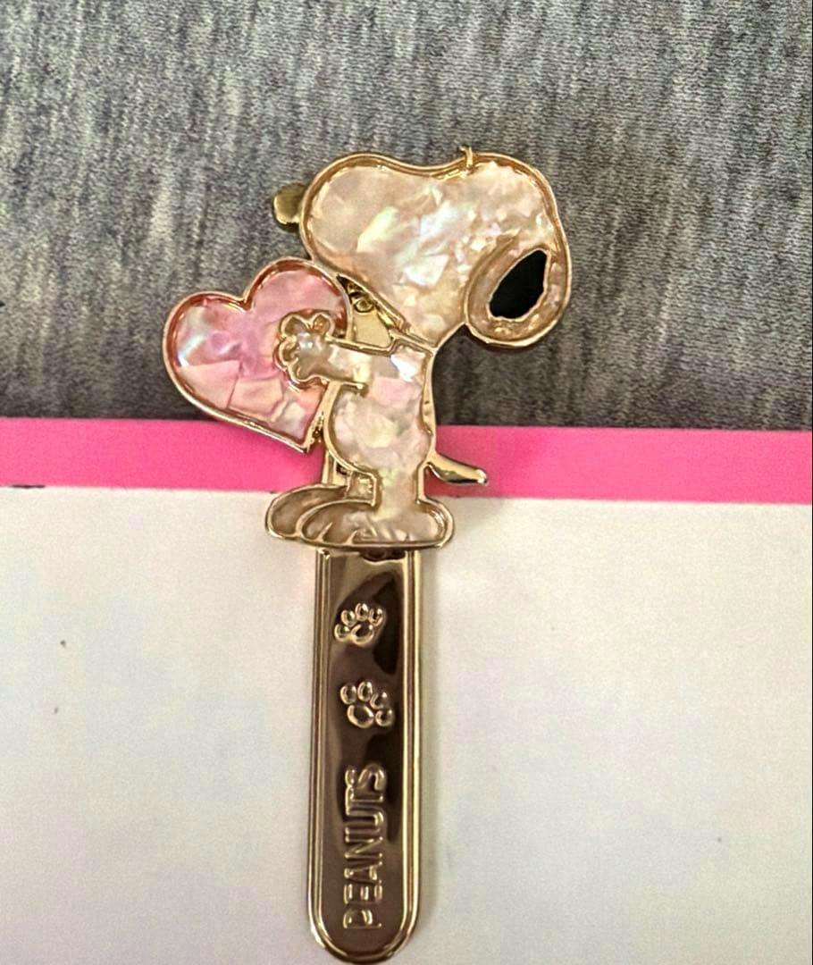 Gold-toned Snoopy bag charm with shell-like inlay holding a heart attached to a metal clip engraved "PEANUTS".