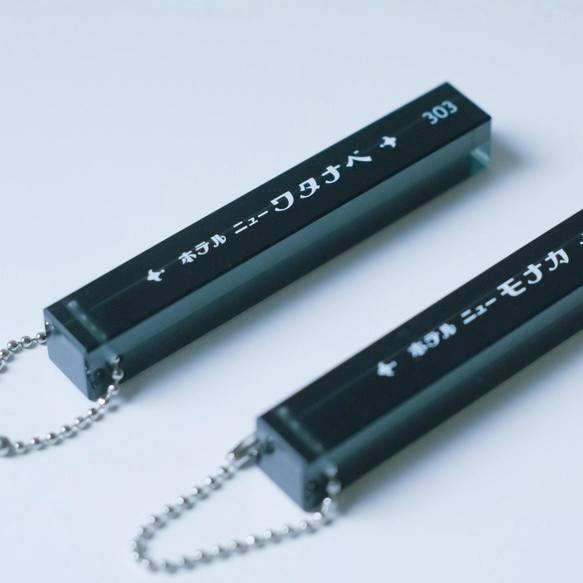 Two black rectangular acrylic hotel room keychains with white Japanese text and metal ball chains on a light background.