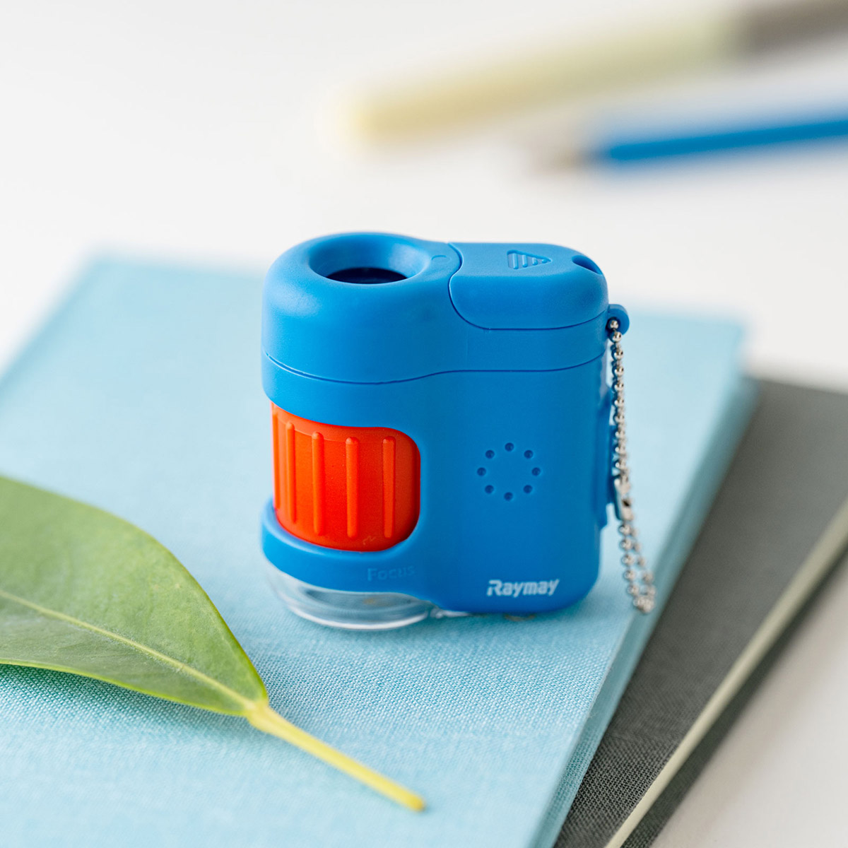 A blue handheld microscope with an orange focusing wheel and attached keychain resting on a notebook.