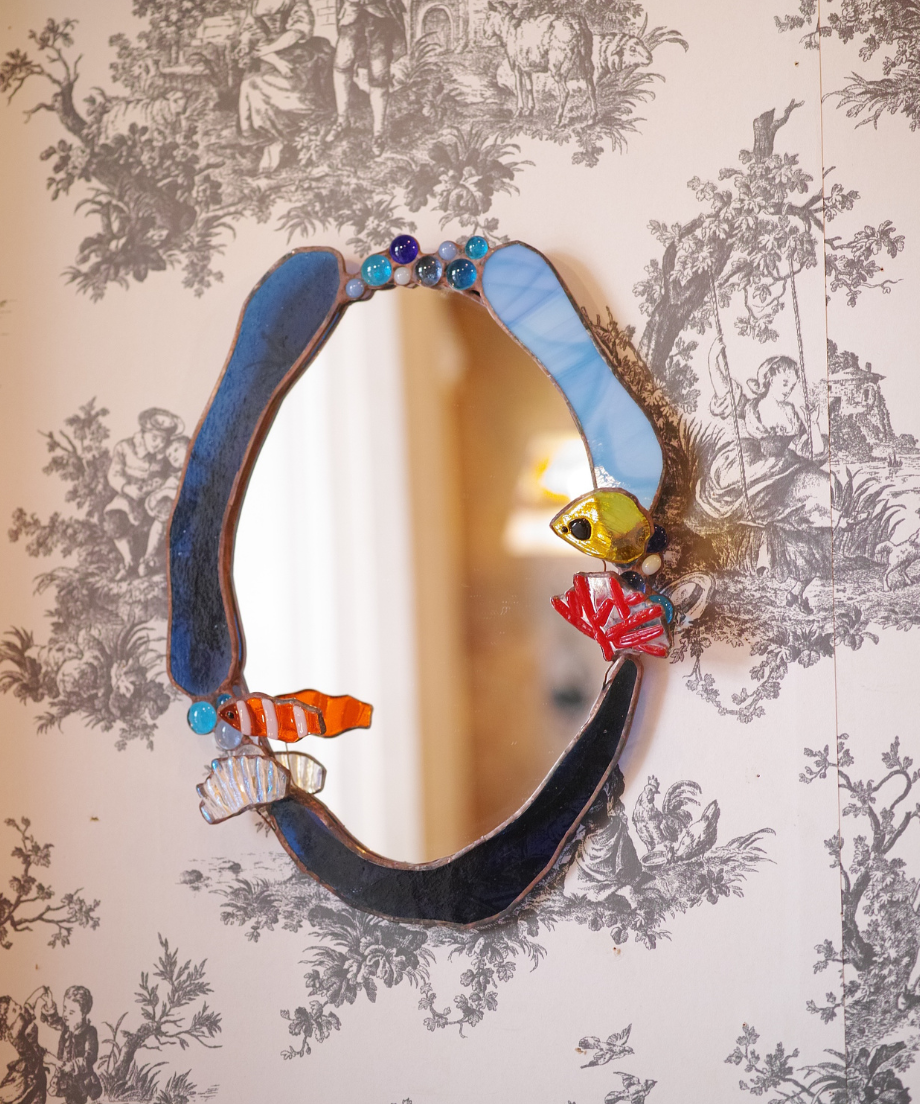 Oval pub mirror with a blue stained-glass frame decorated with small glass fish and coral, shown hanging on patterned wallpaper.