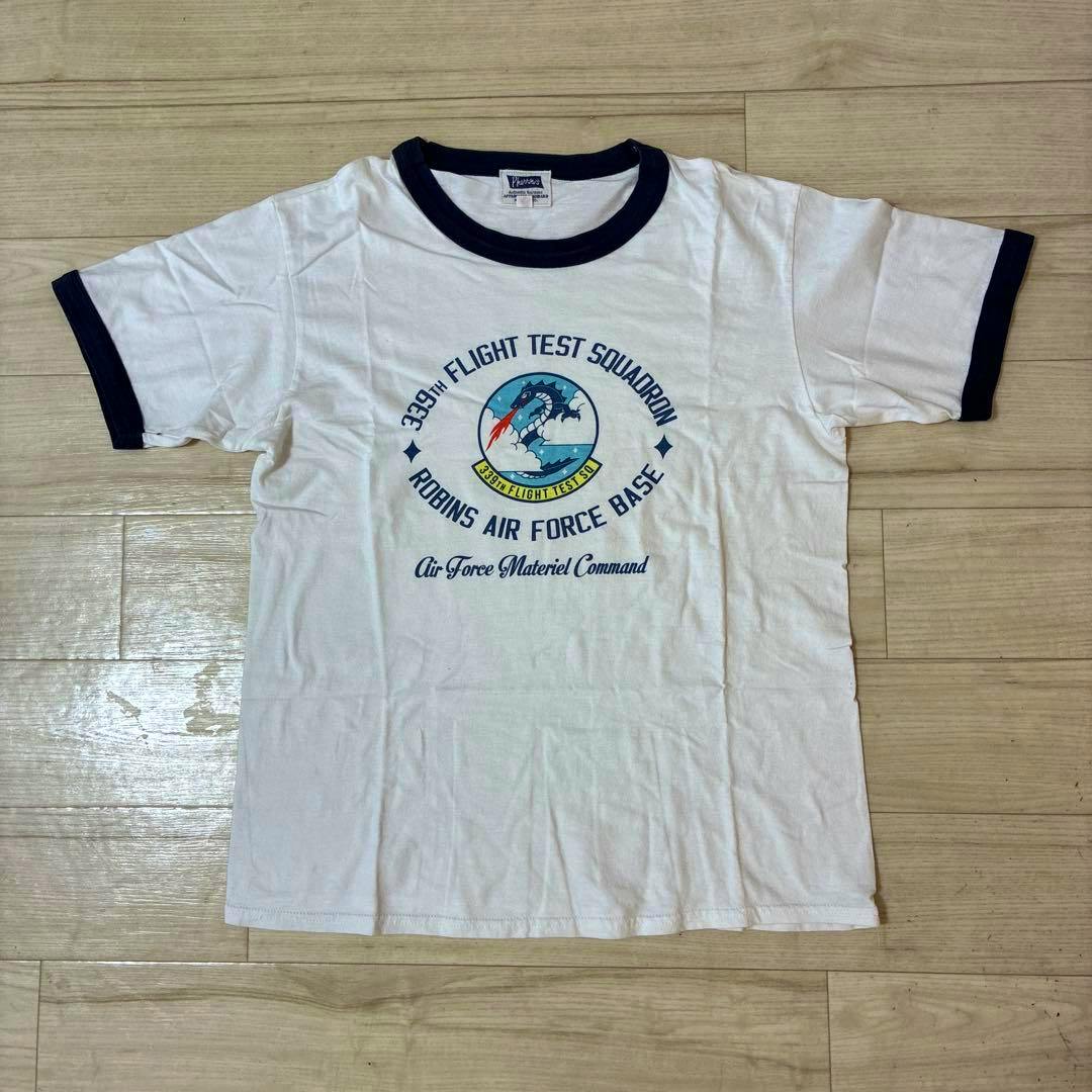 White men’s ringer T-shirt with navy collar and sleeve trim and a 339th Flight Test Squadron / Robins Air Force Base emblem printed on the chest, laid flat on a wood floor.