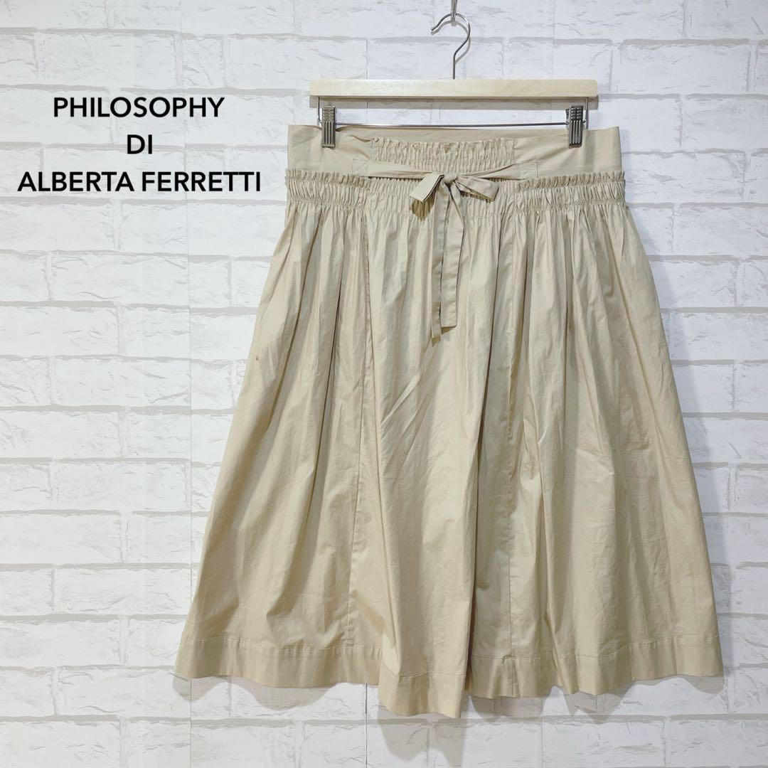 Beige flared skirt with gathered elastic waistband and tied bow hanging on a hanger against a white brick wall.
