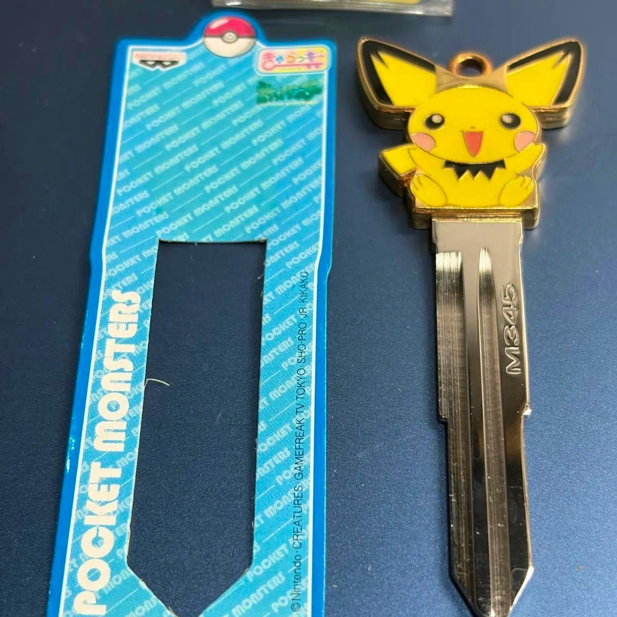 Metal car key blank with a yellow Pichu character top and M345 stamped on the blade, shown next to its blue cardboard backing.