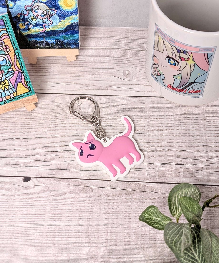 Photograph of the rubber pink cat keychain with metal ring lying on a wooden table beside a mug and a small plant.