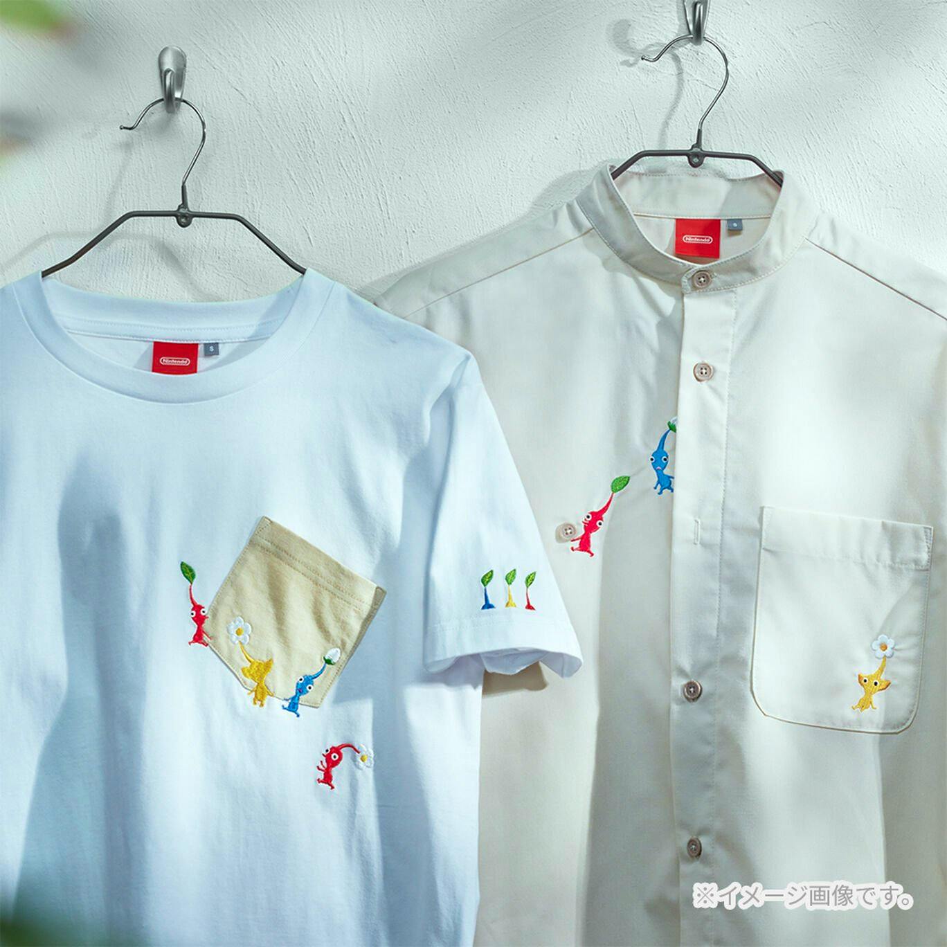Two shirts with Pikmin embroidery hanging on hangers, a white T‑shirt with a beige pocket and a light-colored button-up shirt.