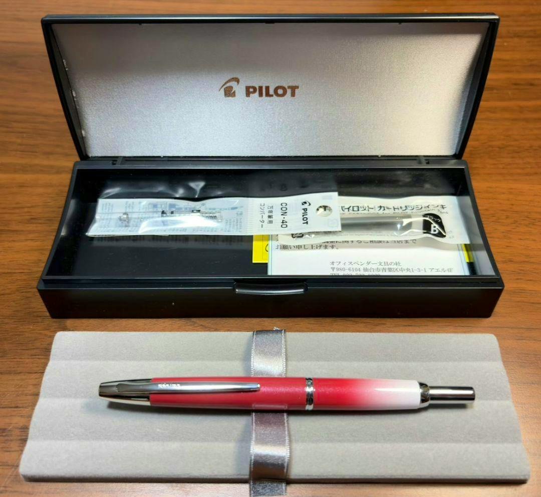 PILOT Capless Decimo fountain pen with red-to-white gradient displayed on a grey pad in front of an open Pilot-branded case with paperwork.