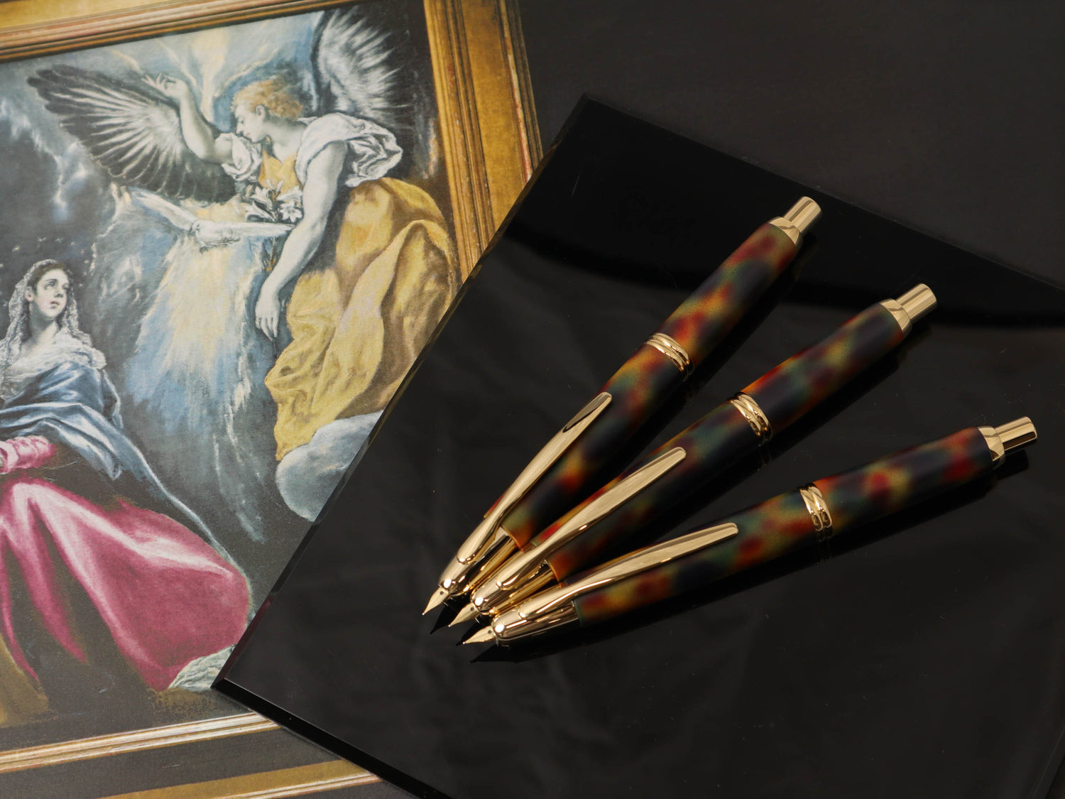 Four capless fountain pens with gold nibs and mottled multicolor barrels displayed on a black surface beside a framed image of El Greco's "Annunciation".