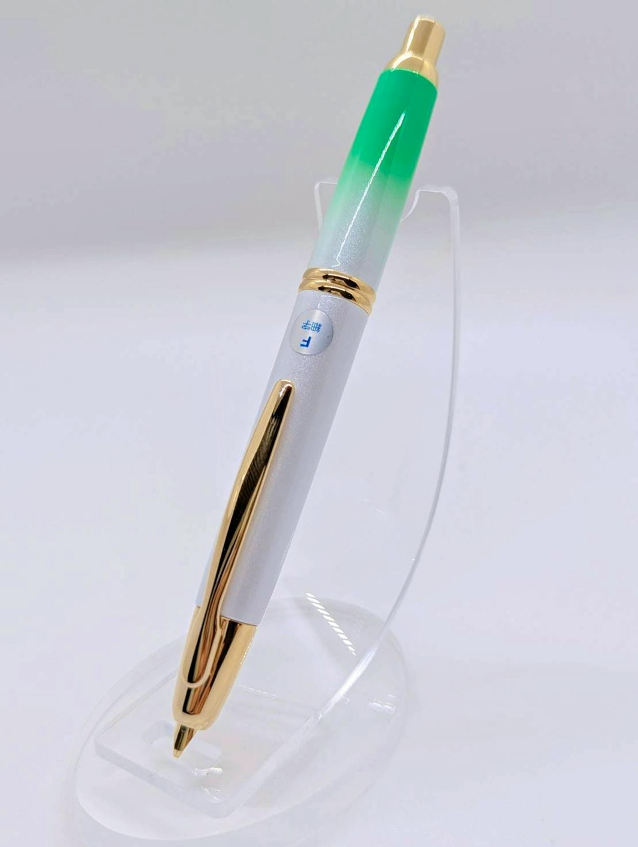 Angled photo of a Pilot Capless fountain pen with a green gradient top, pearlescent white barrel and gold trim displayed on a clear stand.
