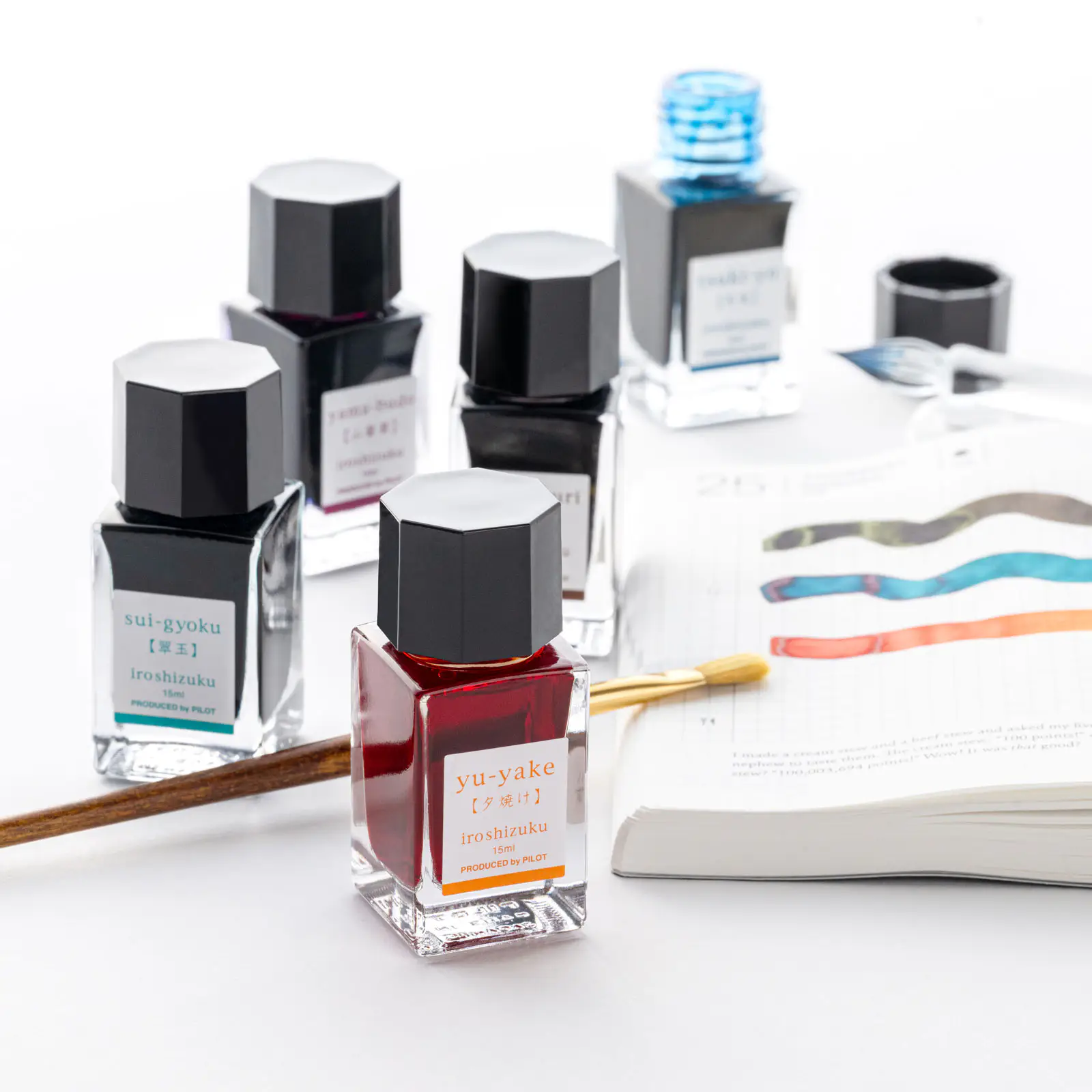 Several Iroshizuku mini fountain pen ink bottles arranged on a white surface next to a notebook and a brush.