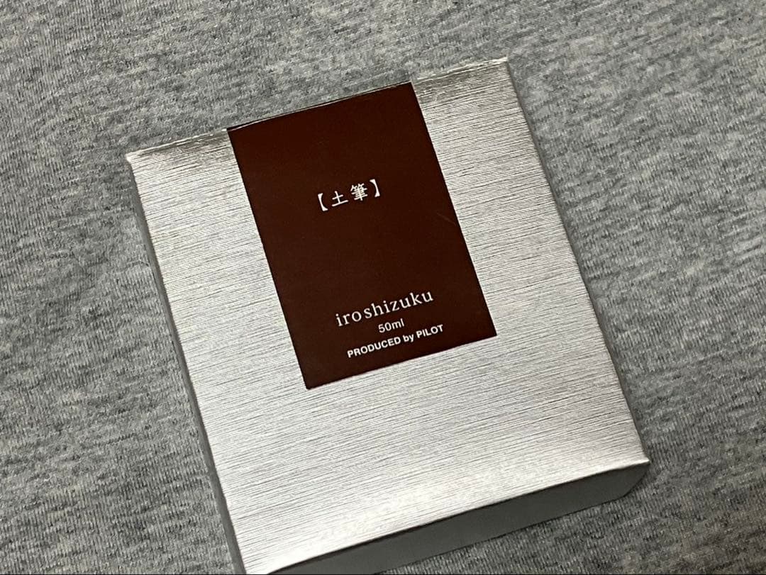 Silver box labeled "iroshizuku 50ml" (土筆) for PILOT fountain pen ink on a gray fabric background.