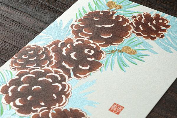 A postcard laid on a wooden surface showing an illustrated pinecone wreath design.