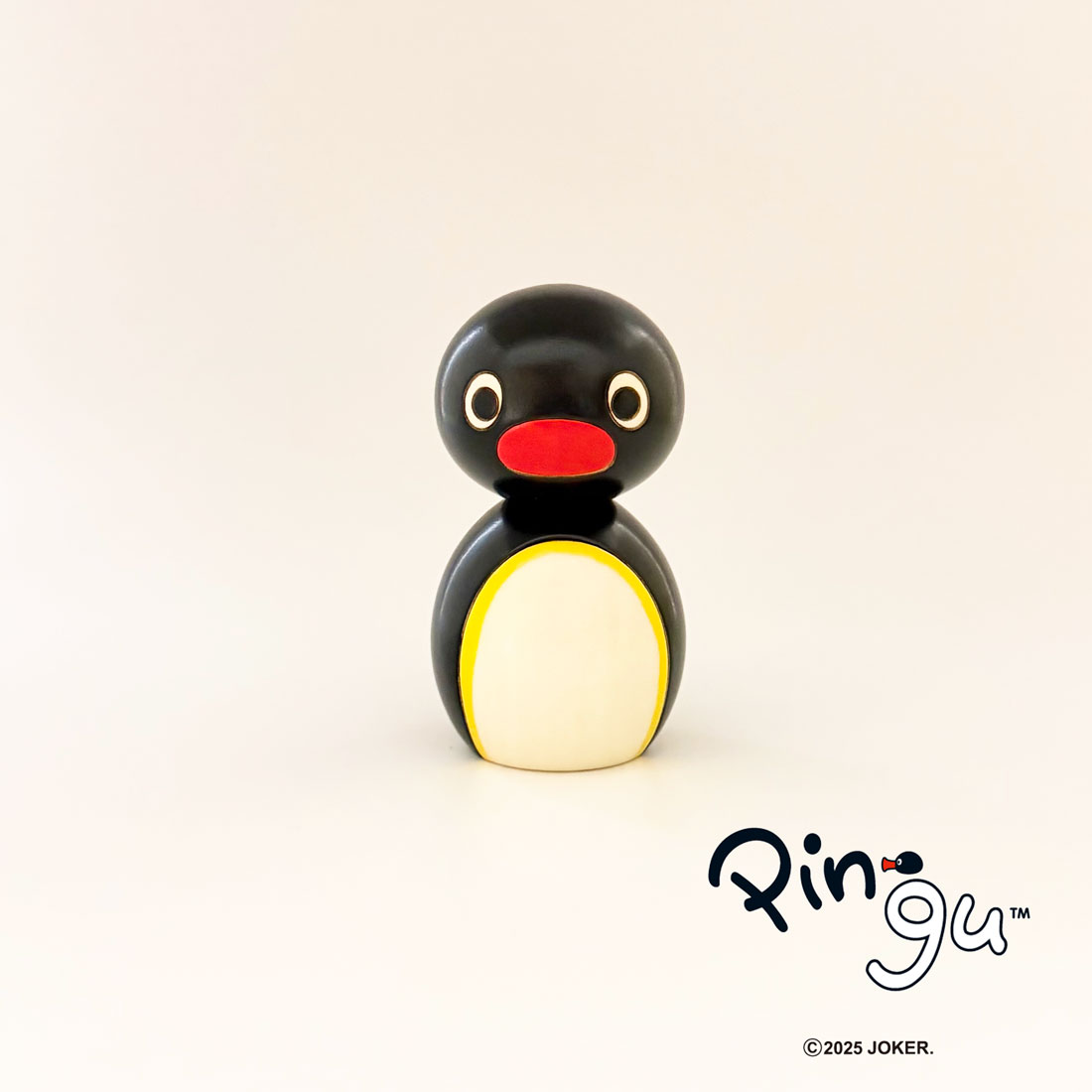 Wooden Pingu kokeshi-style penguin figure with a black head and body, red beak, and white belly with yellow border on a plain background with the Pingu logo.