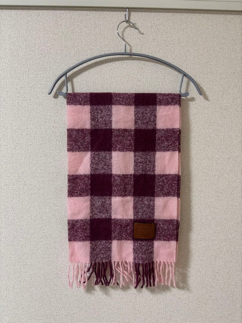 Pink and purple checkered wool muffler with fringed ends hanging on a hanger against a wall.