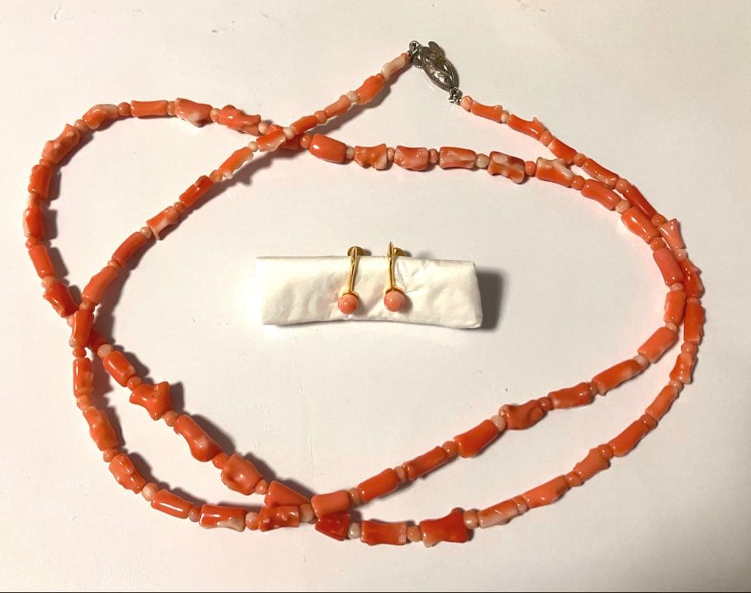 A long pink-red coral bead necklace laid out with a pair of matching small coral drop earrings on a white background.